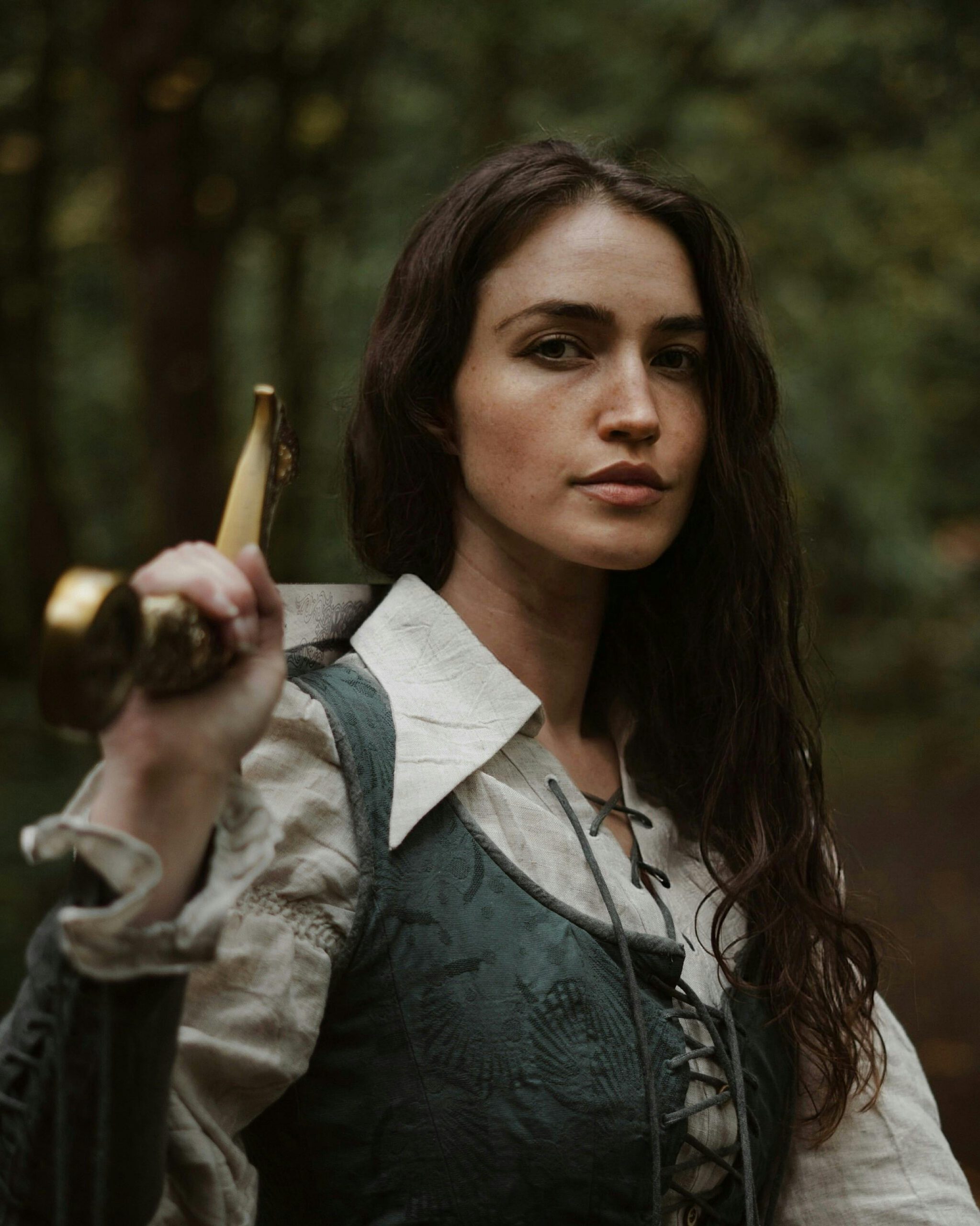 A strong and confident woman in medieval attire poses with a sword in a forest setting.